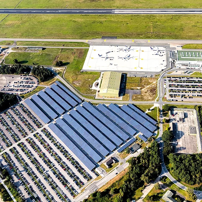 Der Flughafen Weeze aus der Vogelperspektive mit vielen parkenden Autos, einer großen Carportanlage mit Photovoltaikanlage, stehenden Flugzeugen sowie der Start- und Landebahn im Hintergrund.